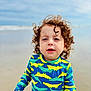 Louie is registered to the contest to win money with this photo: toddler, child, curly_hair, beach, sand, ocean, swimsuit, crocodile_pattern, blue, yellow, green, long_sleeve, water, sky, outdoor, portrait, young_child, casual, daytime, fun