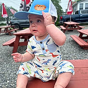 Oliver is registered to the contest to win money with this photo: baby, child, dinosaur_onesie, hat, cap, picnic_table, outdoor, gravel, parked_truck, flags, red_table, cute, sitting, curious, hand_on_hat, summer_clothing, daylight, casual, playful, infant