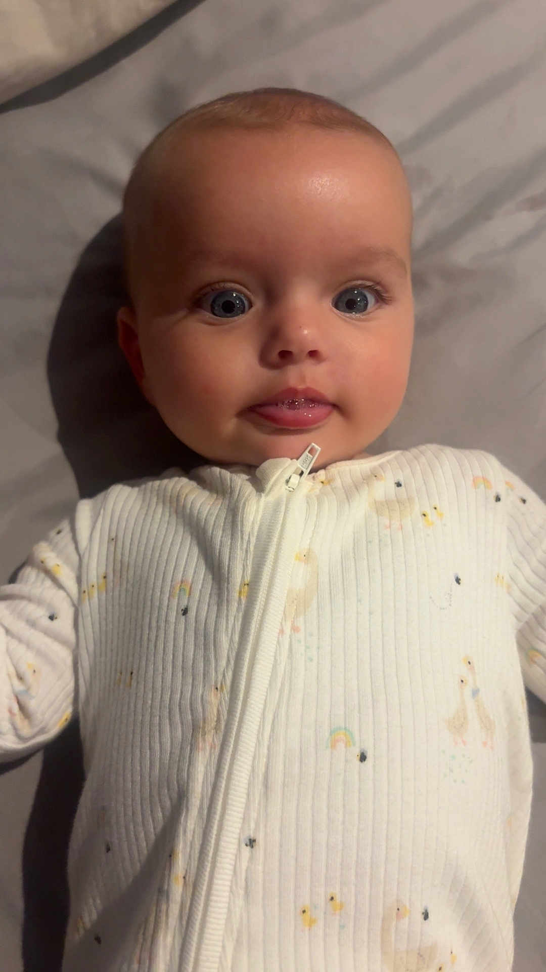 Hallie is registered to the contest to win money with this photo: baby, infant, face, eyes, tongue, onesie, clothing, sleepwear, cute, lying_down, soft_lighting, closeup, child, person, skin, head, expression, bed, fabric, zipped
