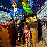 Skyler is registered to the contest to win money with this photo: child, girl, statue, pirate, parrot, amusement_park, ferris_wheel, night, sign, wooden_floor, barrel, colorful, smiling, playland, outdoor, people, casual_clothing, footwear, rope, exhibit