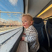 Greyson is registered to the contest to win money with this photo: toddler, child, train, window, snow, rain_drops, curly_hair, striped_shirt, blue_eyes, seat, interior, travel, winter, daylight, landscape, railway_tracks, handrail, curiosity, portrait, candid
