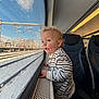 toddler, child, train, window, snow, rain_drops, curly_hair, striped_shirt, blue_eyes, seat, interior, travel, winter, daylight, landscape, railway_tracks, handrail, curiosity, portrait, candid