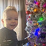 child, toddler, smiling, christmas_tree, ornaments, holiday, indoor, curtains, decorations, lights, festive, happy, person, young_child, celebration, winter, joy, family, seasonal, home