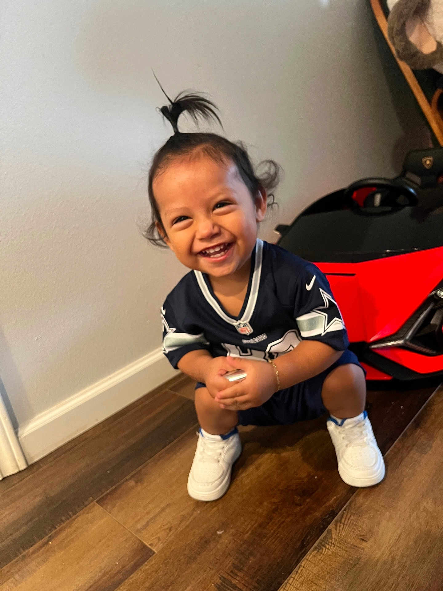 Lucca joined the competition — help win amazing prizes! toddler, child, smiling, squatting, indoor, toy_car, sports_jersey, white_shoes, wooden_floor, happy, cute, hair_ponytail, candid, person, floor, wall, bracelet, young_child, playful, home