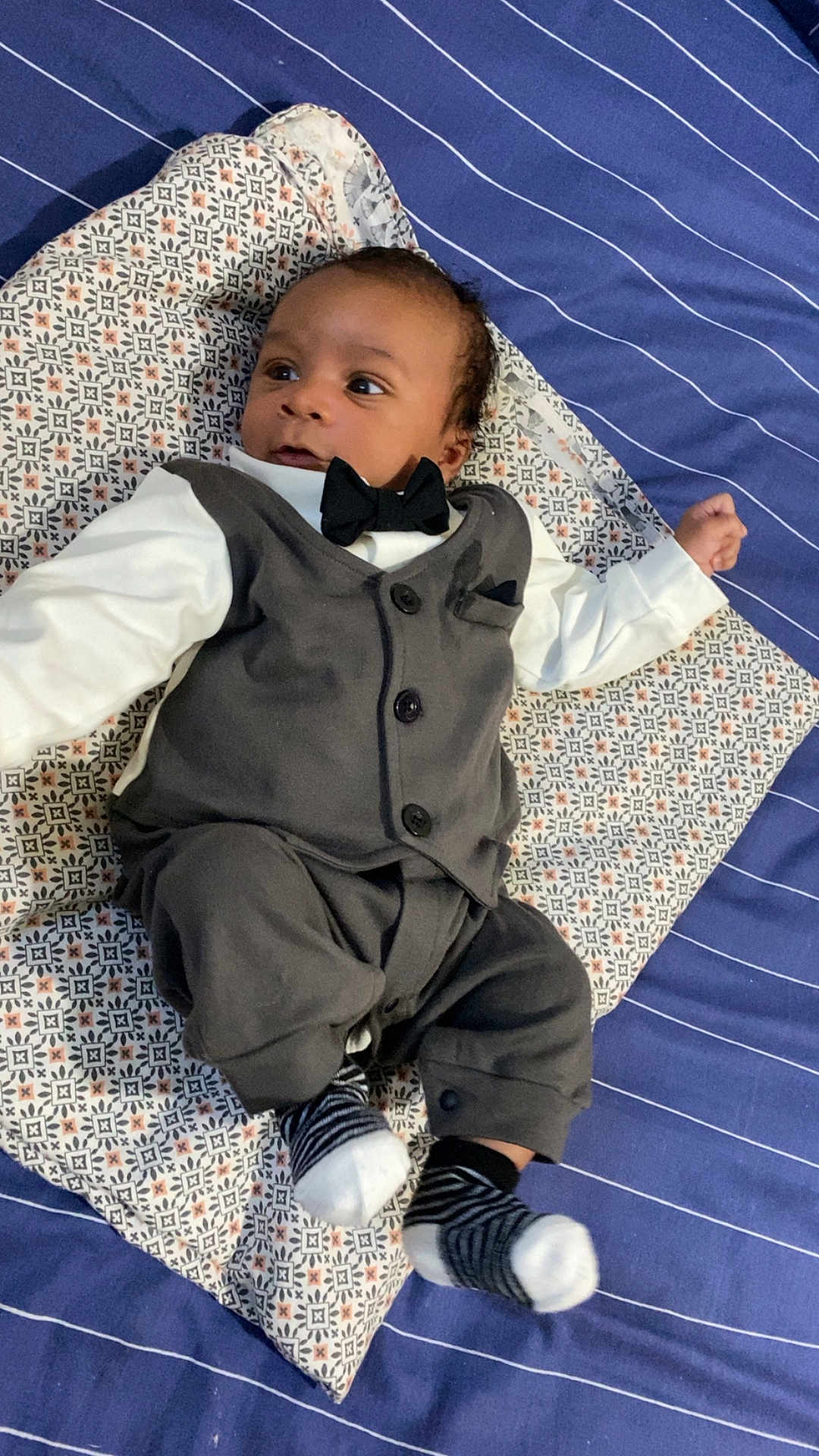 Djeylan participe au concours pour gagner de l'argent avec cette photo : baby, bed, black_hair, bow_tie, clothing, curious_expression, cute, fancy_outfit, indoors, infant, lying_down, patterned_pillow, person, portrait, small_socks, soft_lighting, striped_bedspread, striped_socks, tuxedo, young_child