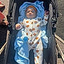 baby, infant, stroller, blanket, pumpkin_pattern, onesie, outdoor, sunlight, footwear, barefoot, sidewalk, person, clothing, headwear, cute, child, baby_clothes, warm, relaxing, daytime