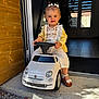 toddler, child, toy_car, white_car, fiat, outdoor, doorway, yellow_cardigan, white_pants, sandals, flower_bows, happy, smiling, cute, playful, steering_wheel, wooden_wall, gravel, sunlight, portrait