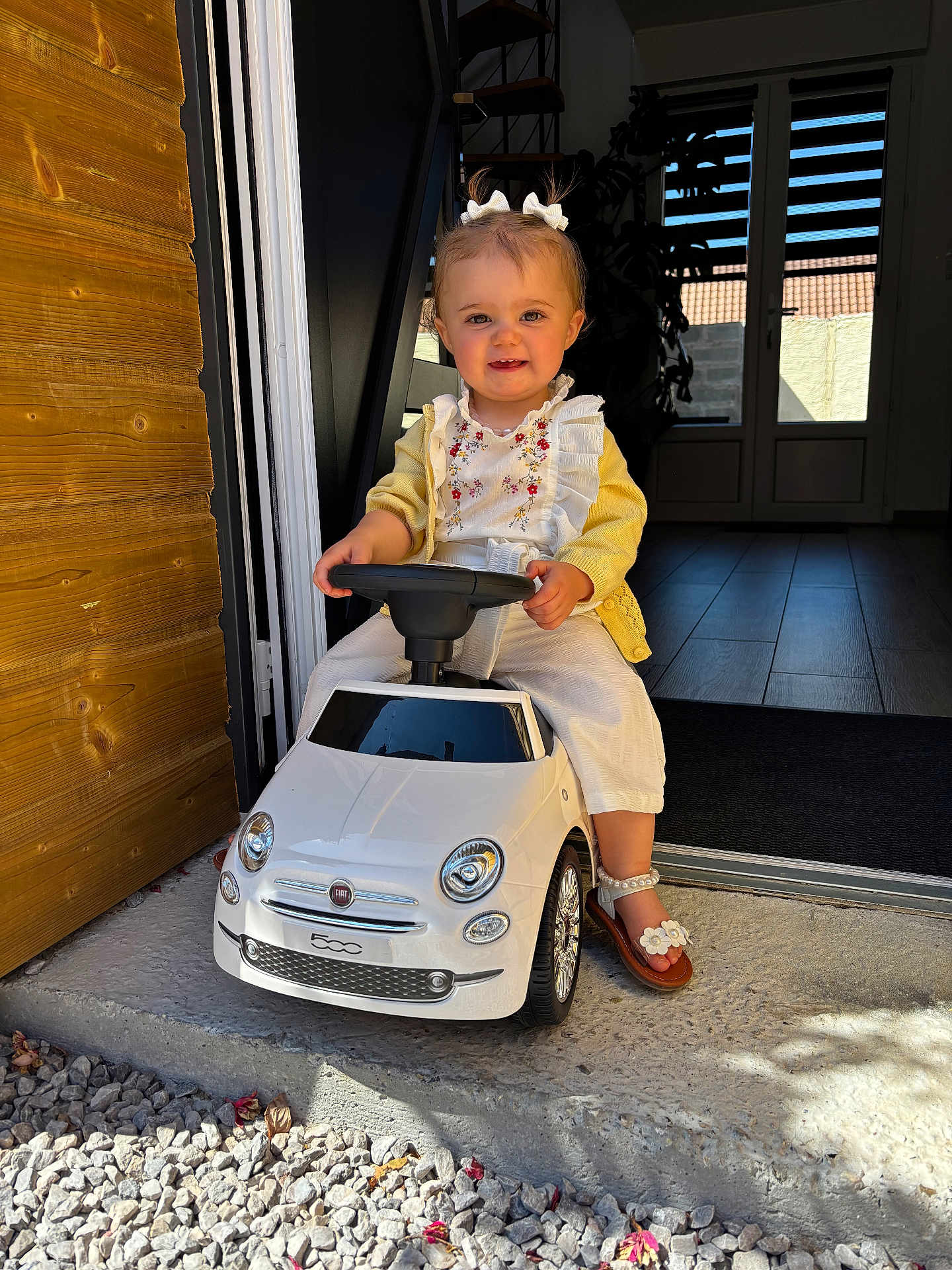 Giulia a rejoint le concours — aidez-le/la à gagner de superbes lots ! toddler, child, toy_car, white_car, fiat, outdoor, doorway, yellow_cardigan, white_pants, sandals, flower_bows, happy, smiling, cute, playful, steering_wheel, wooden_wall, gravel, sunlight, portrait