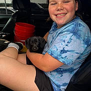 Ollie is registered to the contest to win money with this photo: child, puppy, dog, smile, blue_shirt, tie_dye, car_seat, bucket, black_dog, shorts, shoe, window, vehicle_interior, happy, portrait, casual, young_person, sitting, indoor, pet
