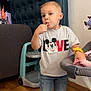 child, toddler, boy, lollipop, tshirt, mickey_mouse, love_text, jeans, sneakers, highchair, baby_seat, wooden_floor, standing, face, eyes, blond_hair, indoors, sofa, furniture, hand