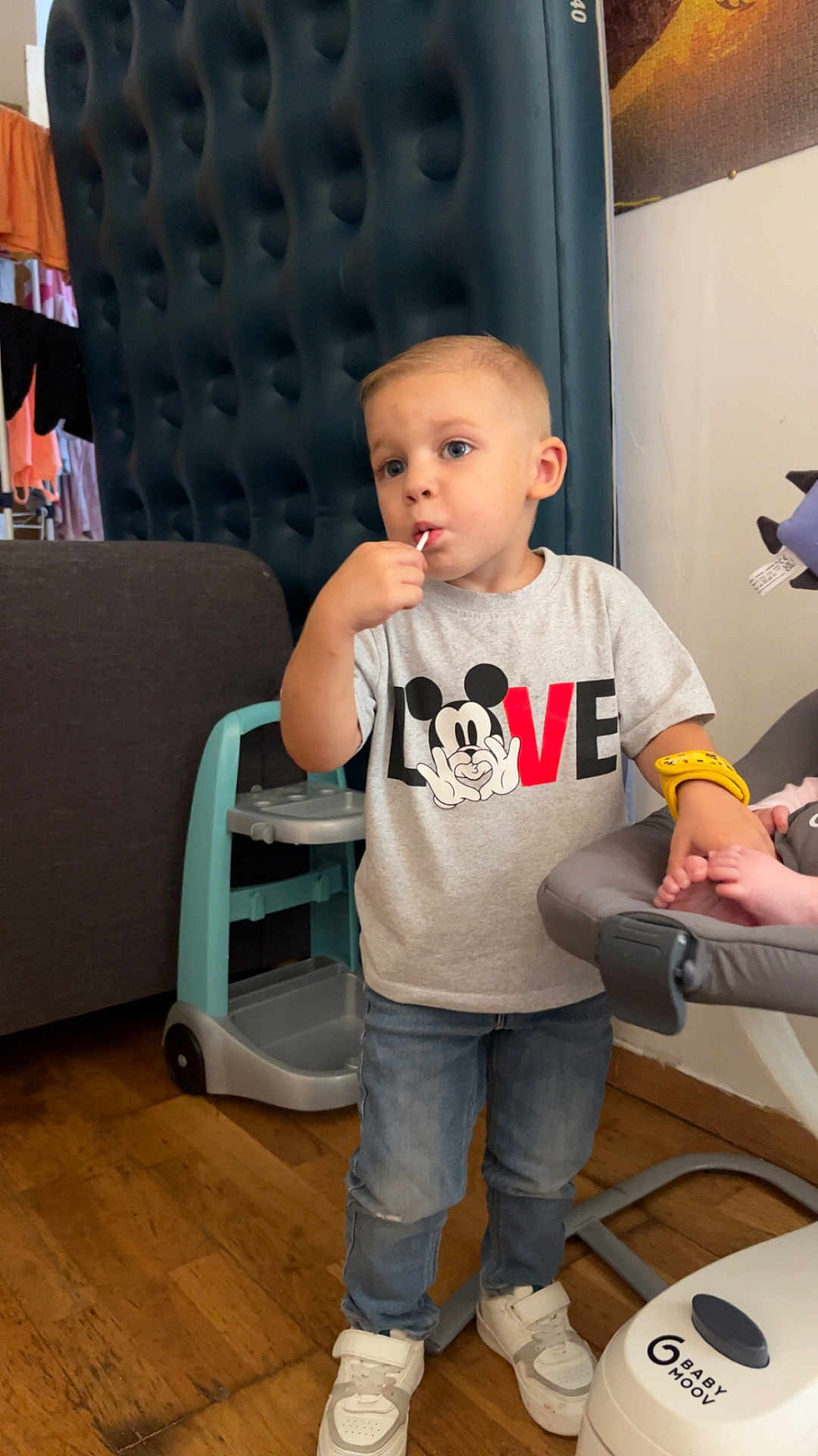 Nolan participe au concours pour gagner de l'argent avec cette photo : child, toddler, boy, lollipop, tshirt, mickey_mouse, love_text, jeans, sneakers, highchair, baby_seat, wooden_floor, standing, face, eyes, blond_hair, indoors, sofa, furniture, hand