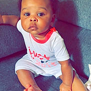 Eleya joined the competition — help win amazing prizes! baby, child, infant, sitting, couch, portrait, indoors, face, eyes, curly_hair, smiling, curious, clothing, onesie, white_outfit, red_trim, bare_feet, furniture, soft_lighting, photography