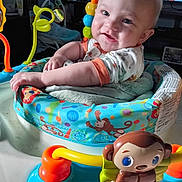 Maverick is registered to the contest to win money with this photo: baby, infant, child, smiling, portrait, indoor, activity_center, jumper, baby_toy, toy_monkey, colorful, playtime, sitting, white_shirt, orange_trim, blue_eyes, blond_hair, happy, television, furniture