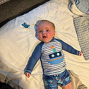 Ralphie is registered to the contest to win money with this photo: baby, child, infant, bed, blanket, pacifier, clothing, swimsuit, face, person, floor, carpet, indoor, smiling, blue, white, pattern, cute, happy, lying