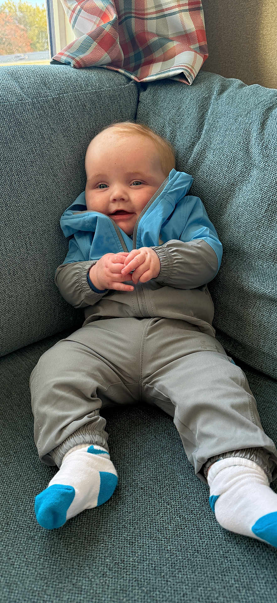 Ralphie joined the competition — help win amazing prizes! baby, child, infant, jacket, blue, gray, socks, white, blue_accent, couch, sofa, plaid_curtain, window, indoor, smiling, hands, young_child, cozy, seated, portrait