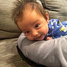 baby, child, infant, face, hair, couch, pillow, blue_clothing, smiling, cute, indoors, person, young_child, lying_down, soft_texture, curious, expression, comfortable, home, relaxed