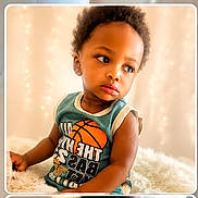 Ethan is registered to the contest to win money with this photo: toddler, child, baby, portrait, sitting, basketball_shirt, curious, serious_expression, brown_skin, curly_hair, face, eyes, lips, indoor, blanket, fluffy_blanket, soft_lighting, string_lights, cozy, photography