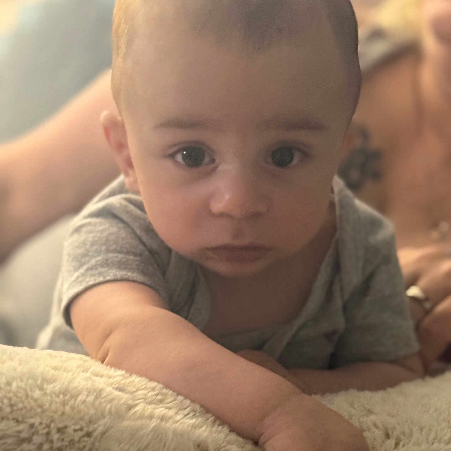 Caiden is registered to the contest to win money with this photo: adult_blurred, arm, baby, blanket, candid, child, close_up, curious, expression, face, grey_shirt, hand, indoor, infant, lying_down, person, portrait, skin, soft_texture, young_child