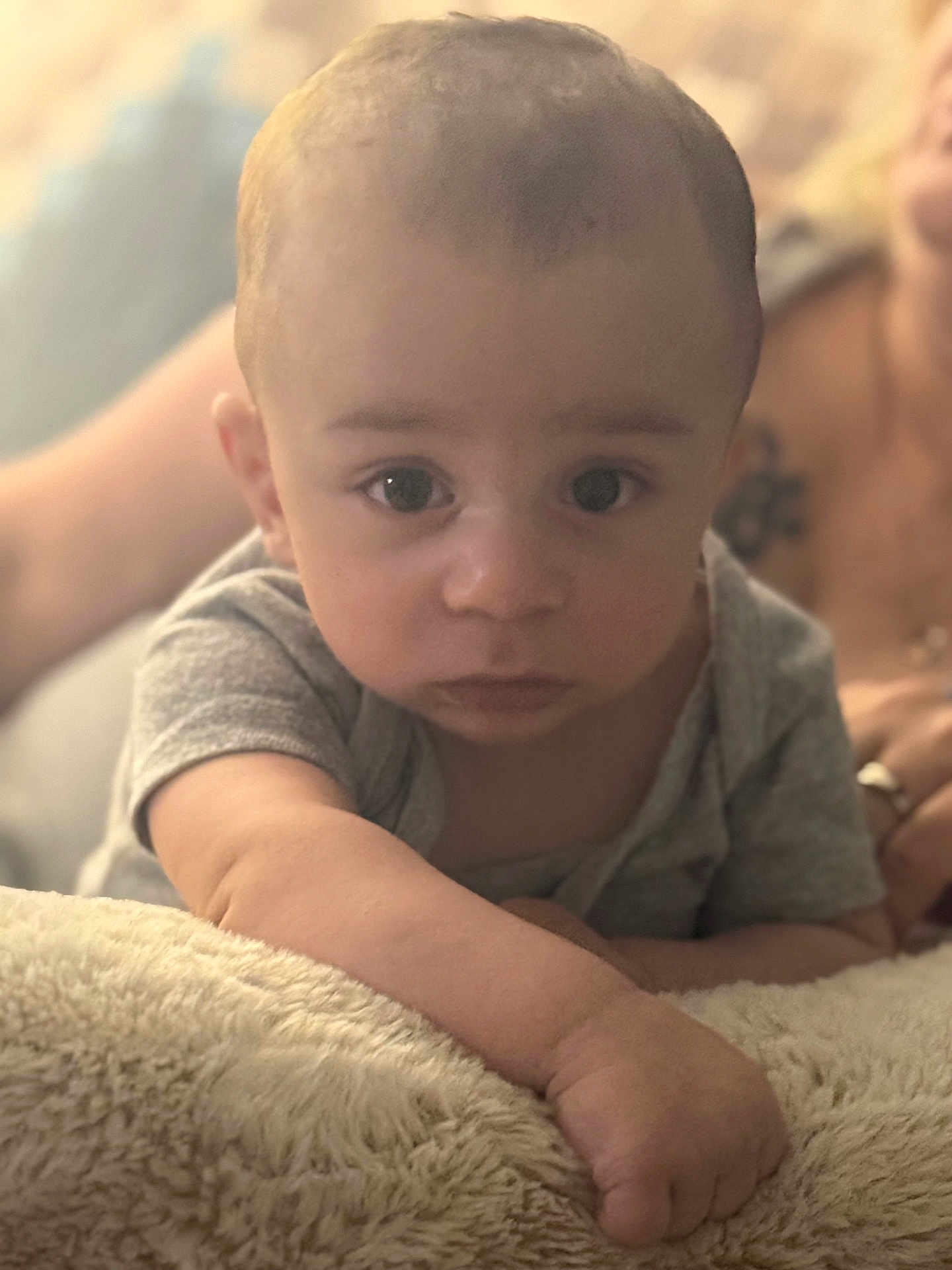 Caiden is registered to the contest to win money with this photo: adult_blurred, arm, baby, blanket, candid, child, close_up, curious, expression, face, grey_shirt, hand, indoor, infant, lying_down, person, portrait, skin, soft_texture, young_child