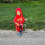 child, kid, hooded_poncho, red_clothing, blue_clogs, grass, greenery, bushes, outdoor, path, sidewalk, nature, person, casual_clothing, daylight, curious_expression, pointing, standing, footwear, young_child