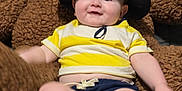 Cameron is registered to the contest to win money with this photo: baby, child, hat, yellow_shirt, blue_shorts, teddy_bear, plush_toy, indoor, smile, cute, infant, sitting, soft_toy, cozy, portrait, person, face, happy, clothing, relaxing