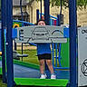 child, playground, play_equipment, blue, green, shorts, sneakers, grass, outdoor, person, park, daylight, fun, curly_hair, panel, car_drawing, legs, standing, smiling, metal_structure