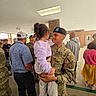 soldier, child, military_uniform, holding, people, indoor, brick_wall, casual_clothing, hat, sweater, pants, shoes, conversation, group, smile, family, portrait, ceiling_light, standing, public_space