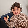 Amelia is registered to the contest to win money with this photo: child, toddler, leopard_print, clothing, potato_chip, snack, chair, indoor, casual, curly_hair, face, person, seated, relaxed, young, cute, expression, light_background, human, portrait
