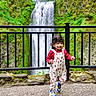 black_rail, child, curly_hair, daylight, floral_overalls, greenery, happy, moss, motion, nature, outdoor, person, rocks, scenic, smiling, sneakers, stone_path, toddler, walking, waterfall