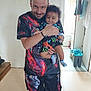 man, baby, child, indoor, smiling, holding, colorful_clothing, socks, shorts, tshirt, floor, doorway, cleaning_supplies, mop, bucket, curly_hair, facial_hair, person, home, casual