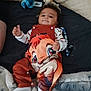 baby, infant, child, bed, blanket, plush_toy, disney, clothing, smiling, thumbs_up, curly_hair, lying_down, cute, comfortable, indoor, soft_toys, person, footwear, striped_sleeves, colorful_outfit