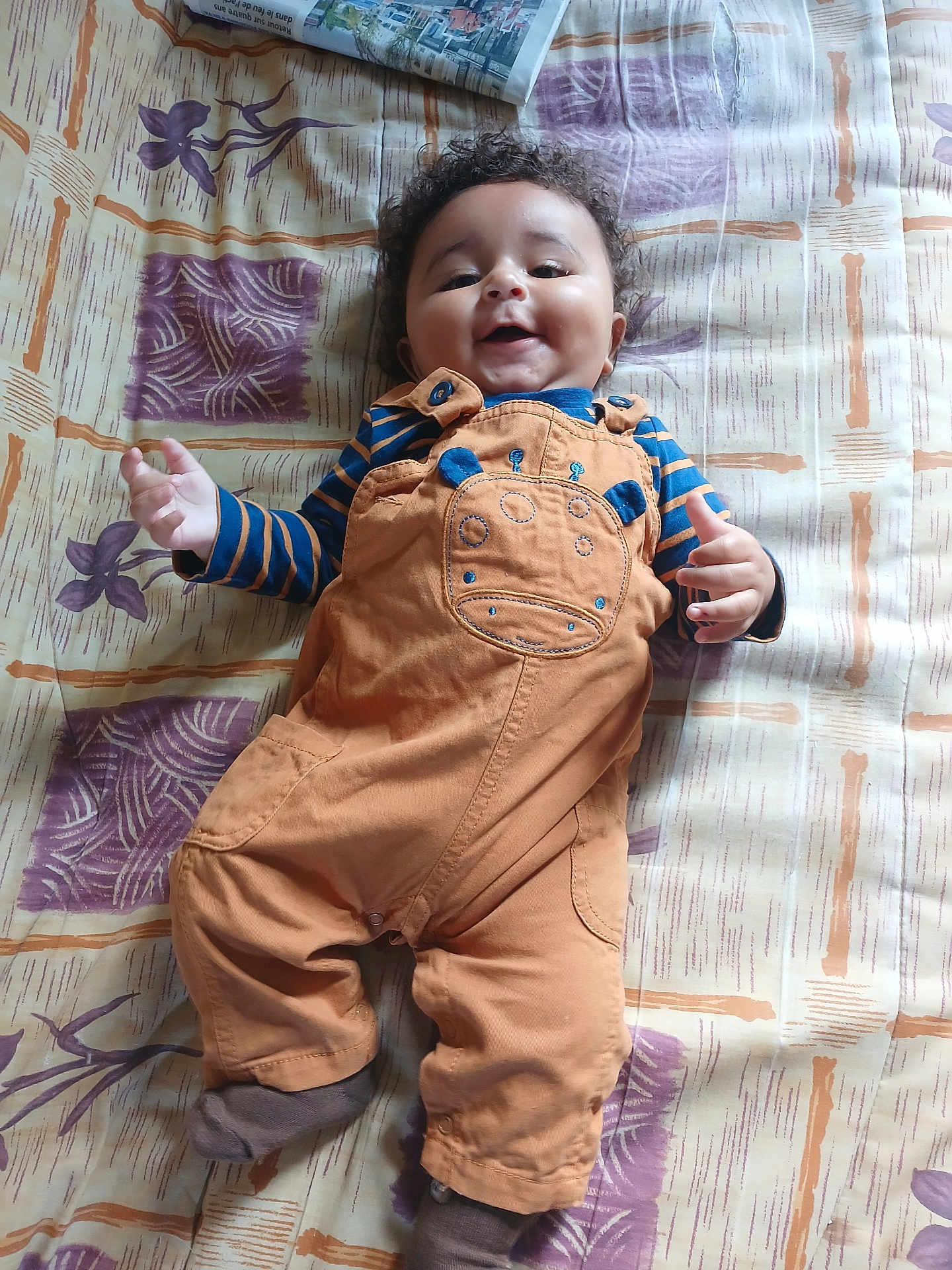 Aaron participe au concours pour gagner de l'argent avec cette photo : baby, child, smiling, curly_hair, overalls, striped_shirt, bedspread, pattern, happy, lying_down, clothing, person, infant, cute, indoor, sock, fabric, face, portrait, playful