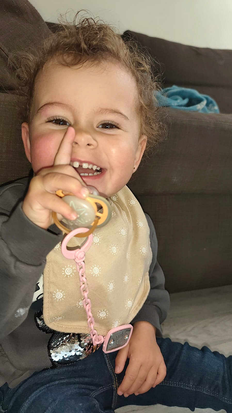 Raquel participe au concours pour gagner de l'argent avec cette photo : toddler, child, smiling, curly_hair, pacifier, bib, casual_clothing, sitting, indoor, couch, happy, playful, hand, face, young_child, jeans, sweatshirt, cute, person, home