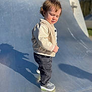 Tenao participe au concours pour gagner de l'argent avec cette photo : toddler, child, outdoor, skate_ramp, shadow, curly_hair, beige_jacket, pants, sneakers, sunlight, daytime, urban, playground, person, standing, casual_clothing, young_child, side_view, expression, curious