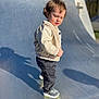 toddler, child, outdoor, skate_ramp, shadow, curly_hair, beige_jacket, pants, sneakers, sunlight, daytime, urban, playground, person, standing, casual_clothing, young_child, side_view, expression, curious