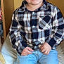 child, boy, smiling, plaid_shirt, blue_jeans, sitting, indoor, chair, happy, casual_clothing, portrait, young, cute, person, furniture, hair, face, hands, light, background