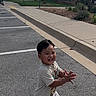 child, toddler, smiling, outdoor, parking_lot, sidewalk, casual_clothing, crocs, happy, clapping, shorts, tshirt, trees, buildings, sunny, daytime, pavement, grass, suburban, person