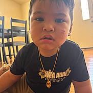 Eziah is registered to the contest to win money with this photo: toddler, child, face, indoor, black_shirt, gold_necklace, scrape, floor, wooden_floor, chair, table, furniture, casual_clothing, portrait, young_child, curious_expression, person, home, daylight, candid