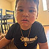 toddler, child, face, indoor, black_shirt, gold_necklace, scrape, floor, wooden_floor, chair, table, furniture, casual_clothing, portrait, young_child, curious_expression, person, home, daylight, candid