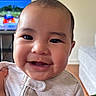 Lovella is registered to the contest to win money with this photo: baby, smiling, face, infant, onesie, clothing, indoor, person, child, cute, happy, portrait, closeup, head, skin, cheeks, hand, furniture, television, couch