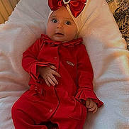 Renesmee is registered to the contest to win money with this photo: baby, infant, red_clothing, headwear, bow, cushion, white_blanket, indoors, portrait, wide_eyes, festive, cute, child, soft_texture, hand, seat, expression, person, cozy, comfortable