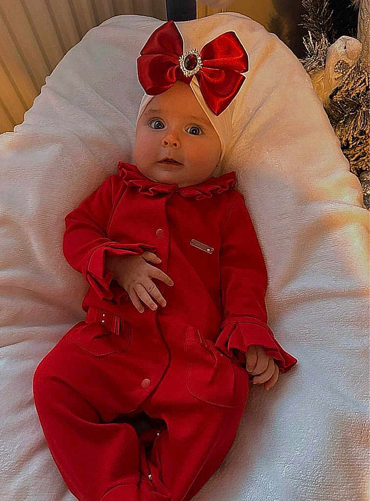 Renesmee is registered to the contest to win money with this photo: baby, infant, red_clothing, headwear, bow, cushion, white_blanket, indoors, portrait, wide_eyes, festive, cute, child, soft_texture, hand, seat, expression, person, cozy, comfortable