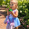 toddler, child, outdoor, sunny, greenery, flowers, pretzel, ball, blue_tutu, pink_purse, stuffed_animal, hair_tied, cute, walking_path, nature, daylight, shoes, playful, garden, portrait