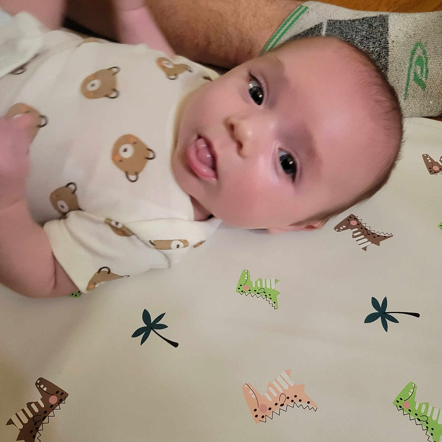 Sonny is registered to the contest to win money with this photo: baby, infant, child, lying_down, portrait, face, eyes, tongue, wide_eyed, onesie, clothing, dinosaur_pattern, play_mat, indoor, wooden_floor, adult, arm, leg, close_up, candid
