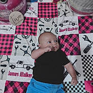 James is registered to the contest to win money with this photo: baby, infant, child, black_shirt, blue_pants, patchwork_quilt, tools_pattern, truck_pattern, lying_down, curious_expression, indoor, fabric, colorful, cute, young_child, portrait, clothing, person, floor, soft_texture