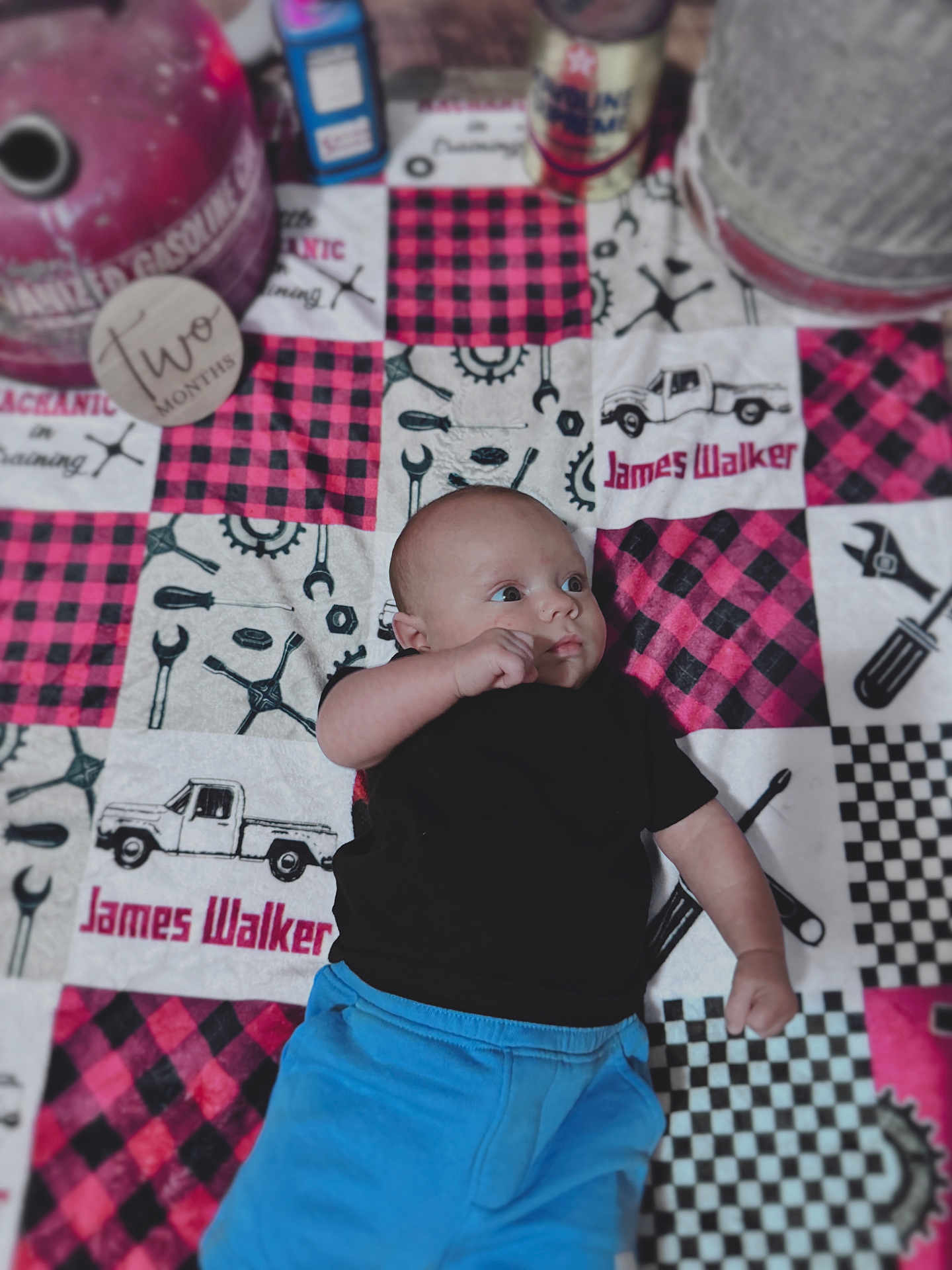 James is registered to the contest to win money with this photo: baby, infant, child, black_shirt, blue_pants, patchwork_quilt, tools_pattern, truck_pattern, lying_down, curious_expression, indoor, fabric, colorful, cute, young_child, portrait, clothing, person, floor, soft_texture