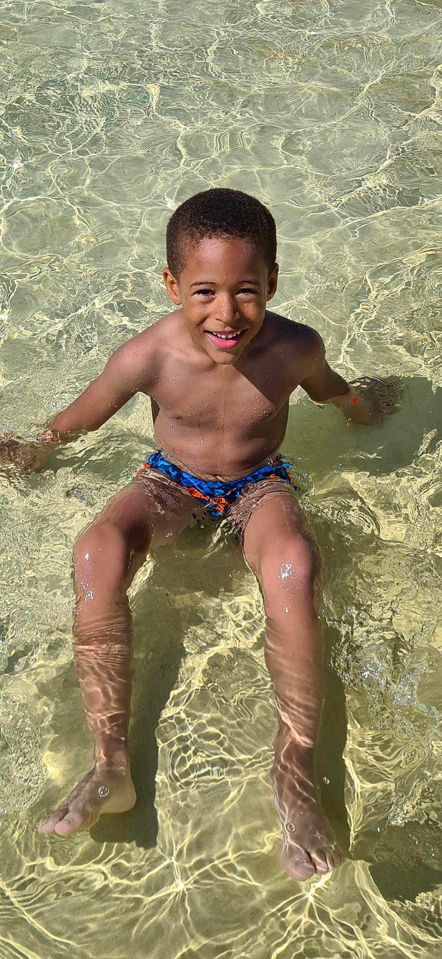 Sohann participe au concours pour gagner de l'argent avec cette photo : child, boy, water, swimming_trunks, sitting, smiling, sunlight, summer, playful, wet_skin, clear_water, outdoor, recreation, happy, young, skin, legs, feet, ripples, joy