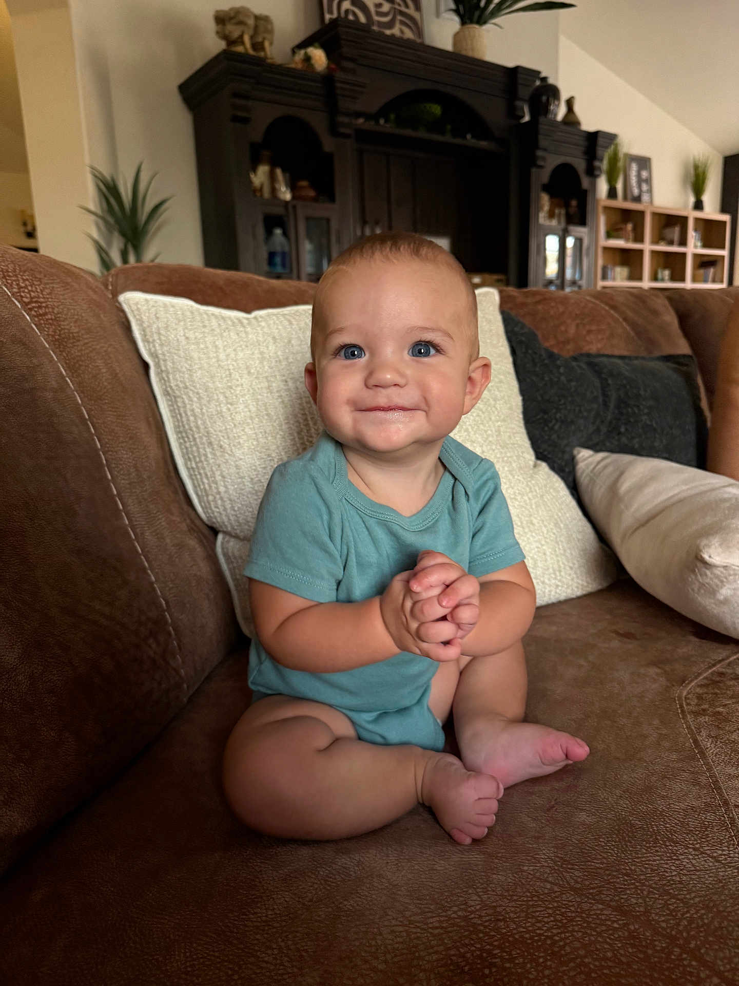 Jesse is registered to the contest to win money with this photo: baby, child, blue_eyes, smiling, sitting, cross_legged, hands_clasped, brown_couch, leather_couch, pillows, indoor, living_room, furniture, cabinet, plant, homedecor, cute, infant, portrait, happy