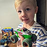 child, boy, toddler, portrait, smiling, toy, monster_truck, toy_vehicle, playtime, striped_shirt, blond_hair, indoors, home, wooden_floor, colorful_toys, closeup, happy, holding_toy, face, photography