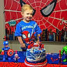 child, birthday, birthday_cake, spiderman, superhero, captain_america, hulk, action_figures, party, celebration, portrait, smiling, blond_hair, blue_shirt, indoors, decorations, spider_web, table, colorful, cake